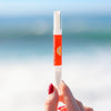A hand holding a tube of FRSHN UP Lightening Whitening Pen - Original Formula - 2-pack sunscreen lip balm against a blurred beach background.