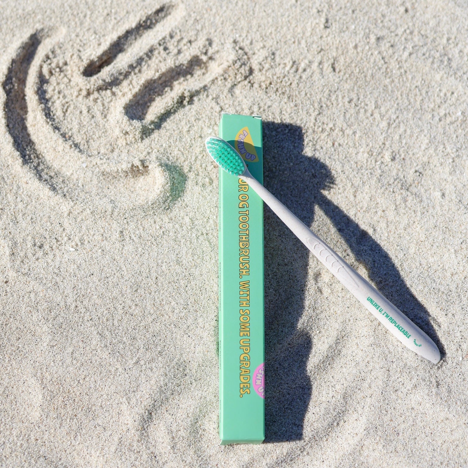 A FRSHN UP Ultimate Lightening Whitening Vault toothbrush lying on sand next to its packaging with a sun design drawn in the background, embodying the essence of oral care.