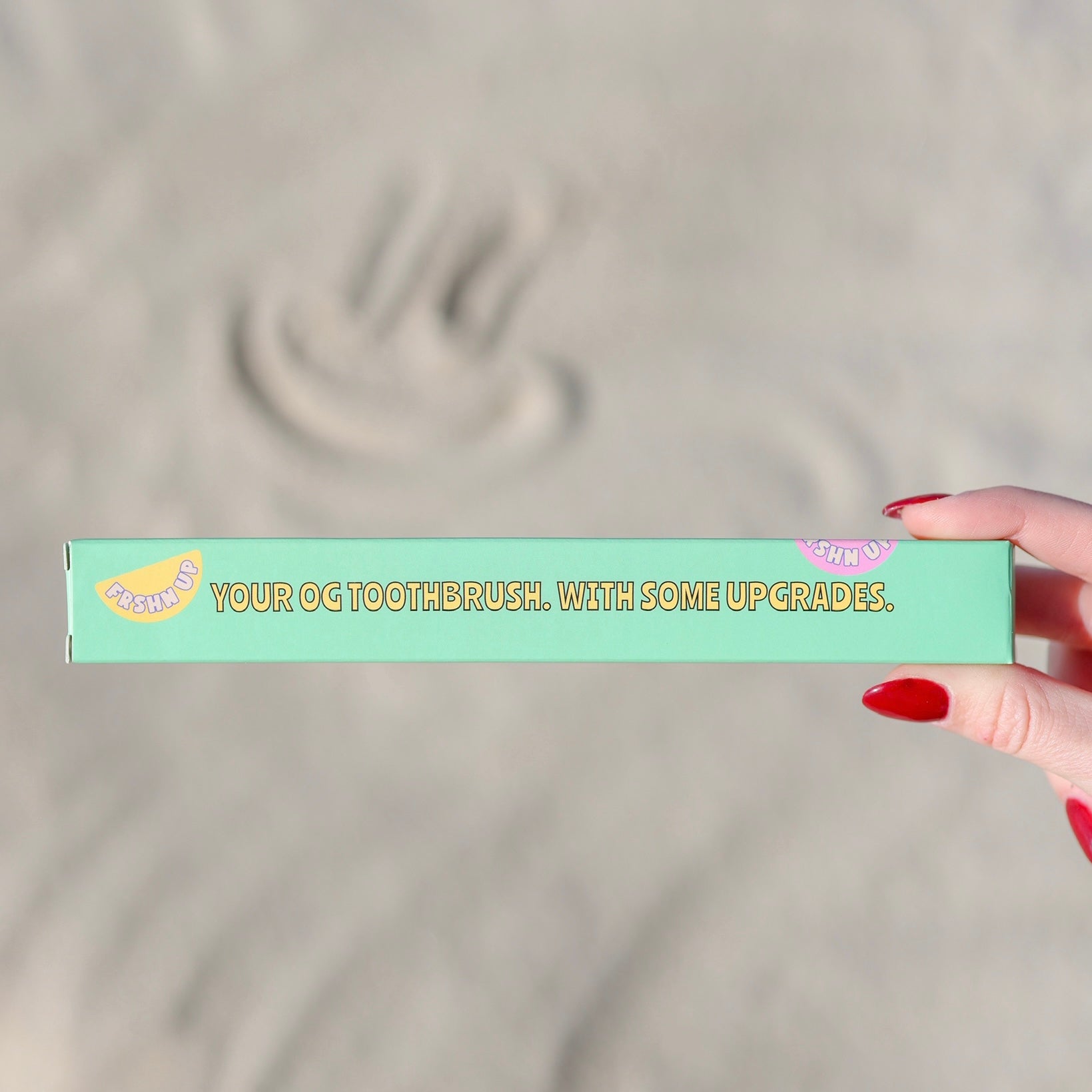 A hand holding an FRSHN UP eco-friendly toothbrush box with the text "your OG Toothbrush. With some upgrades.