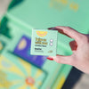 A hand with red nails holds a box of Whitening Toothpaste Tabs labeled "FRSHN UP: Take us with you." The green, eco-friendly package mentions the benefit "Good for: traveling, gym, at home." In the background, the packaging is partially visible.