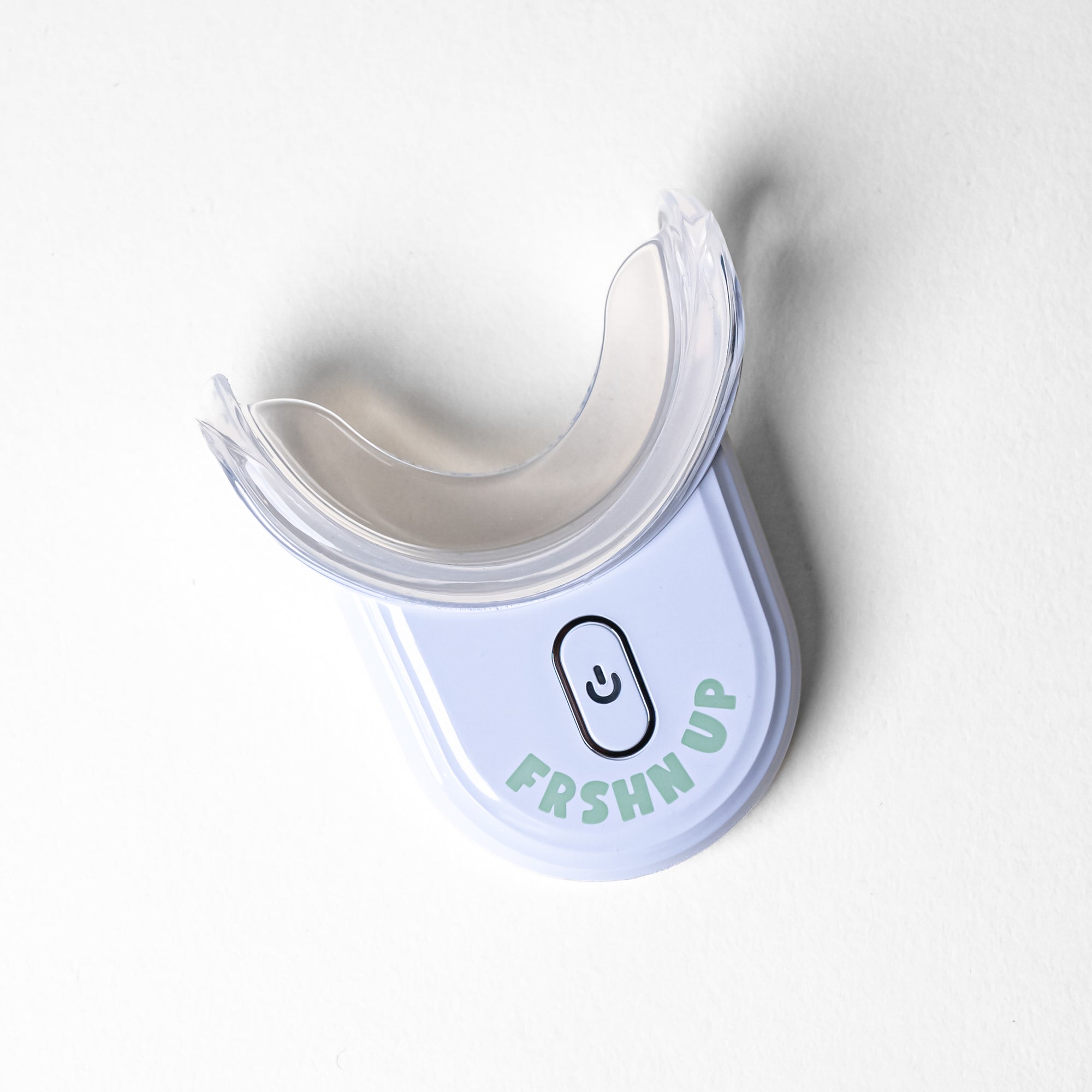 A white and clear dental mouthpiece device with a button and the words "FRSHN UP" printed on the front, designed with a teeth whitening hydrogen-peroxide formula endorsed by dental professionals, the Deluxe Lightening Whitening Kit by FRSHN UP.