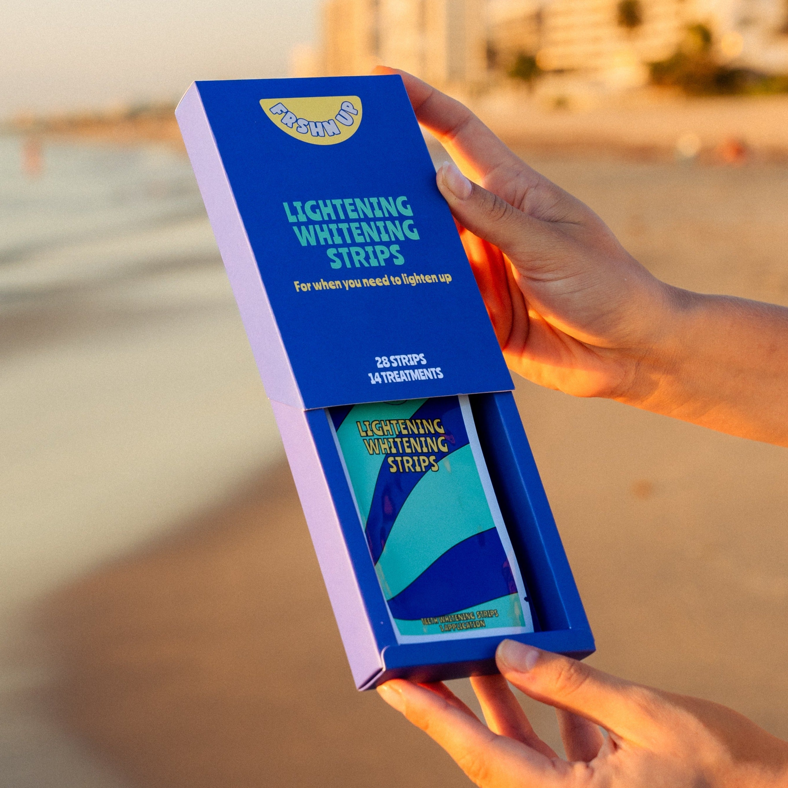 A woman holding up a blue box of FRSHN UP sensitivity-free Lightening Whitening Strips on the beach.